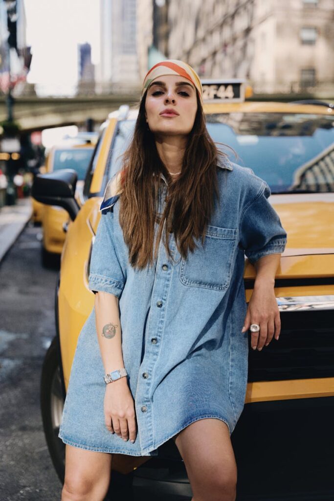 Trendy woman in denim dress poses confidently in urban New York City with a yellow taxi backdrop.