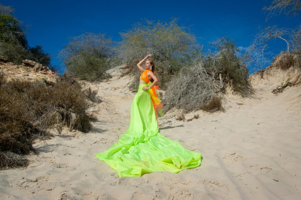 Stylish woman in neon dress poses on sandy dunes under clear blue sky.