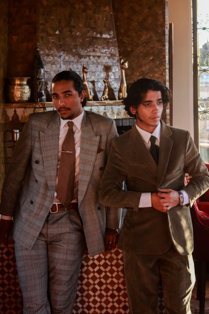 Two men dressed in vintage suits pose elegantly indoors against a stylish backdrop.