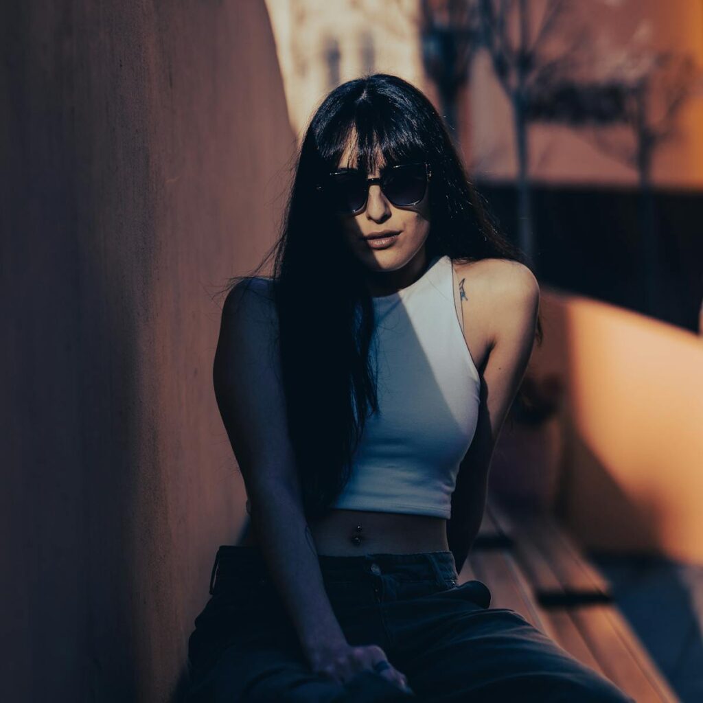 Fashionable young woman in sunglasses sitting outdoors in strong sunlight shadows.