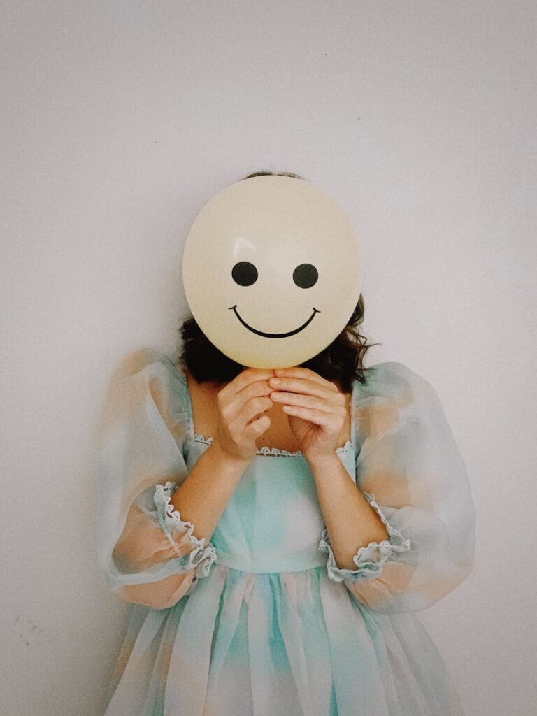 Elegant woman in a pastel dress holding a smiley face balloon against a simple background.