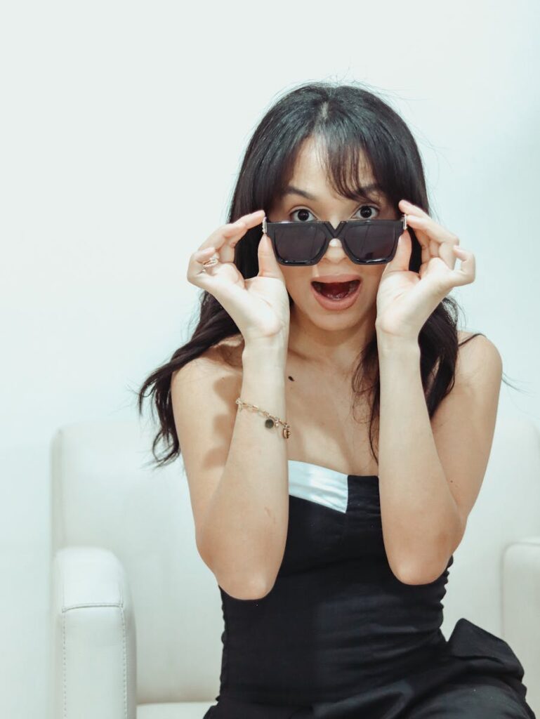 Woman in black dress looking surprised while lifting sunglasses indoors.
