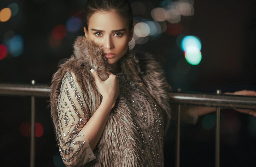 Stylish woman posing in a fur coat against a bokeh night backdrop.