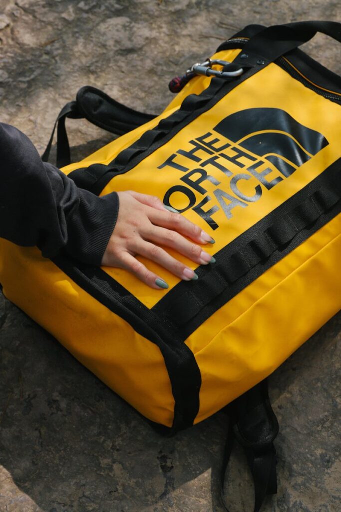 A bright yellow North Face backpack on a stone surface with a person's hand resting on it.