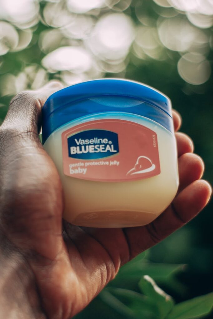 A close-up of a hand holding a jar of Vaseline Blue Seal against a blurred background.