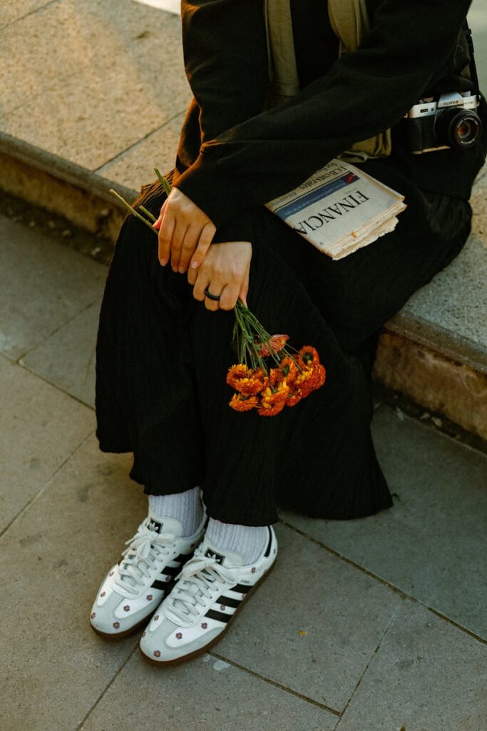 A person sits on a bench holding flowers and a newspaper, wearing casual attire.