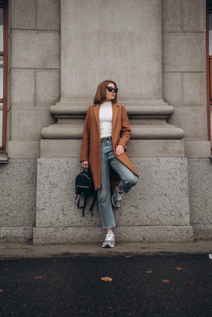 Woman in a brown coat and jeans leaning against a stone wall in an urban environment.
