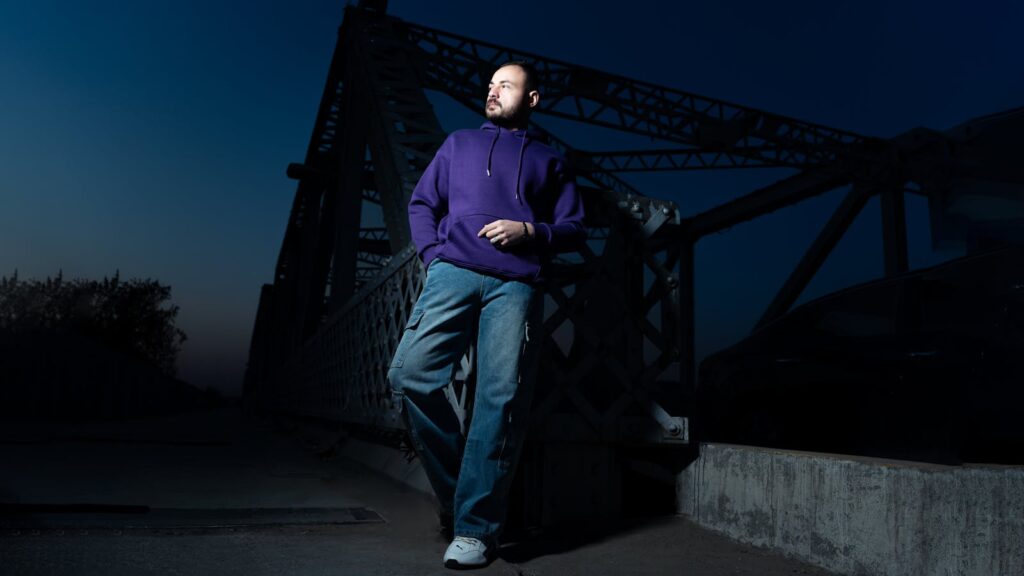 Man in casual attire poses confidently on a steel bridge at dusk.