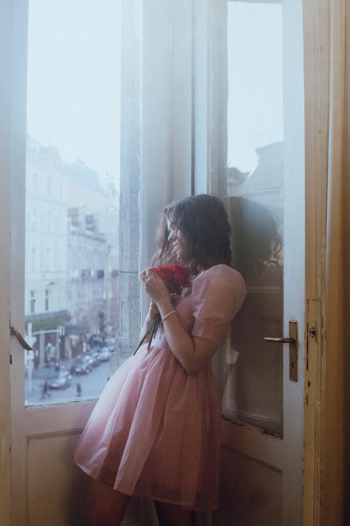 A woman in a pink dress holds flowers by a large window overlooking a city street.