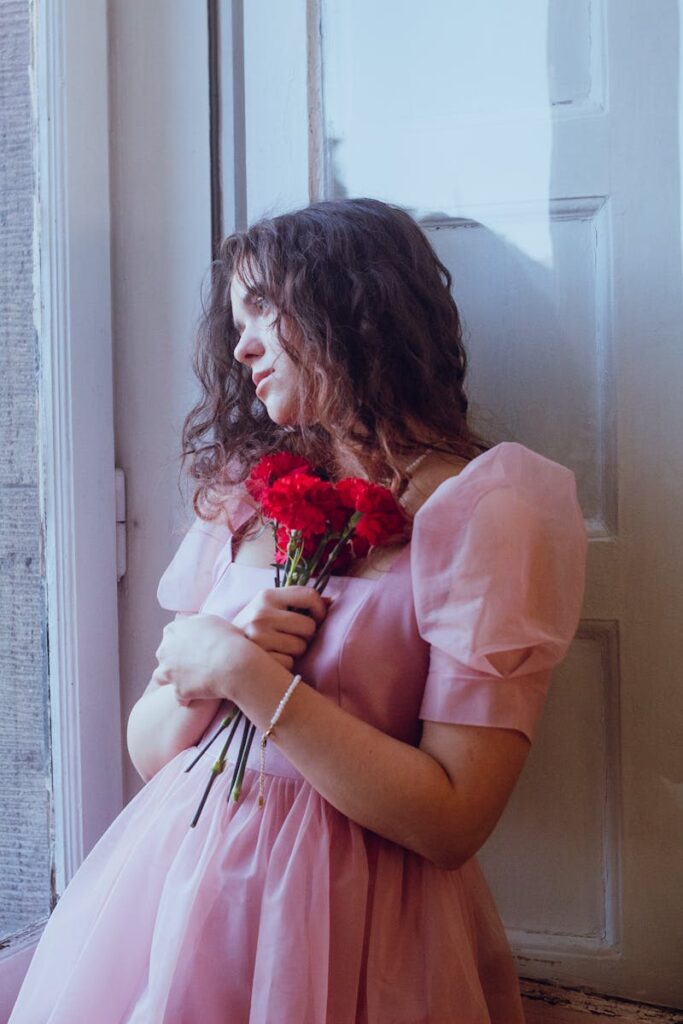A woman in a pink dress holds red flowers, gazing out a window.