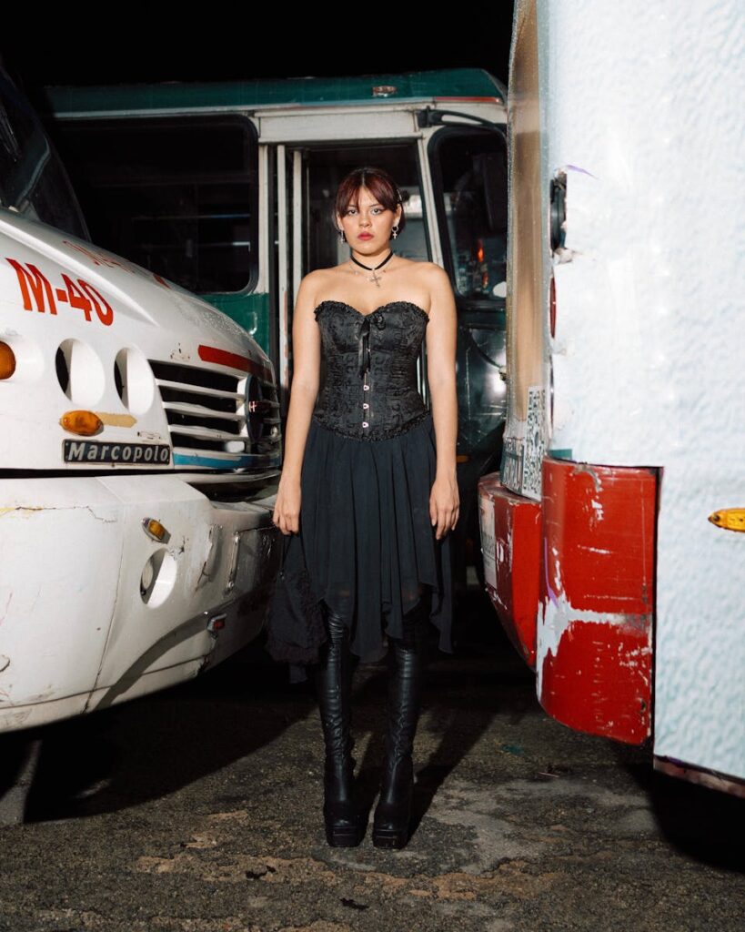 A woman in gothic attire stands between two buses at night, exuding mystery and style.
