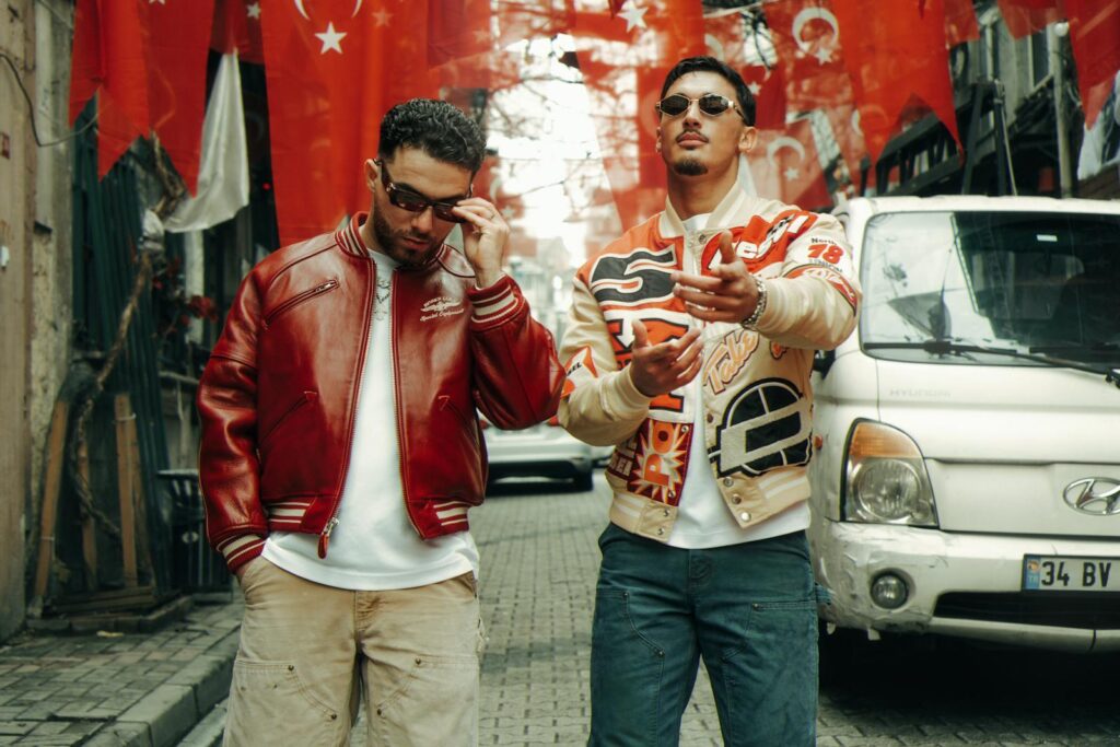 Two fashionable men on city street with red flags hanging, showcasing urban style.
