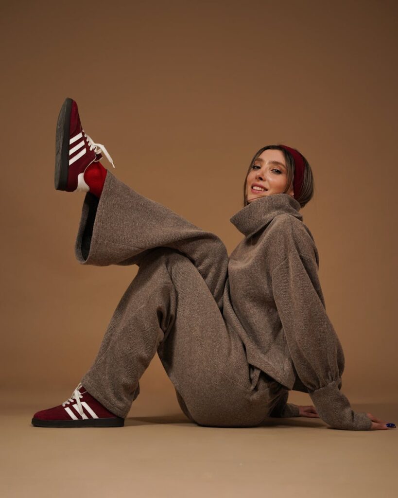 Fashionable woman in brown outfit and red sneakers posing playfully on a brown backdrop.