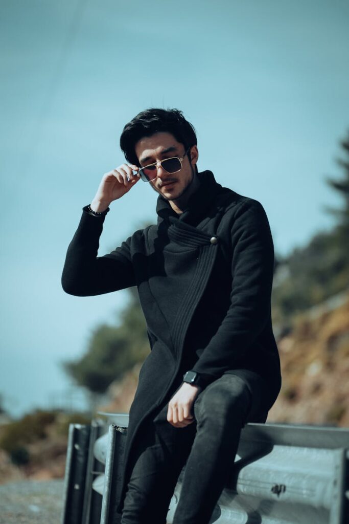 A fashionable young man in a black coat and sunglasses enjoying an outdoor setting