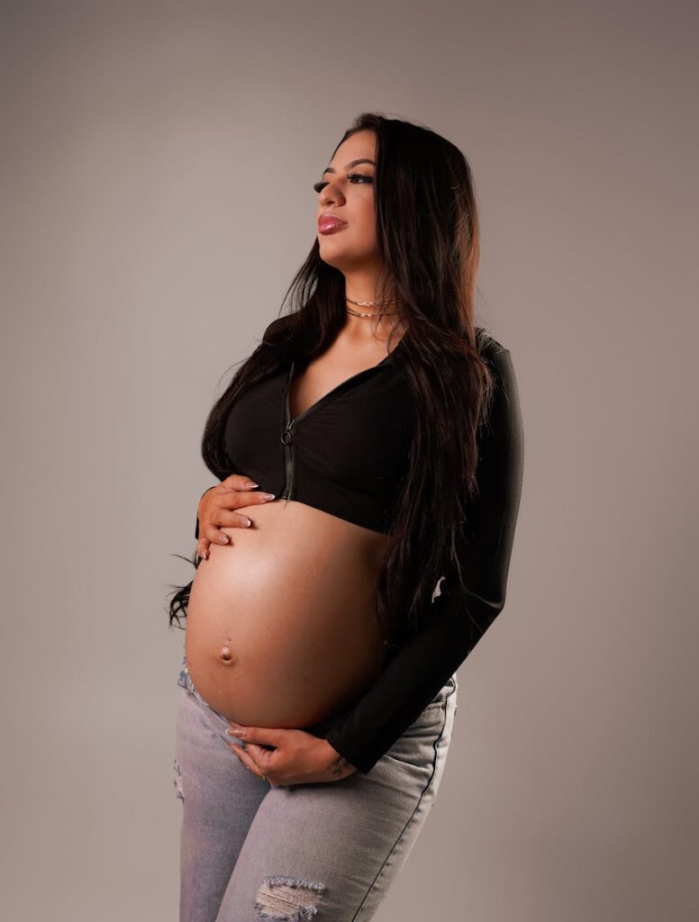 Pregnant woman holding her belly, dressed in casual clothing, standing against a neutral background.