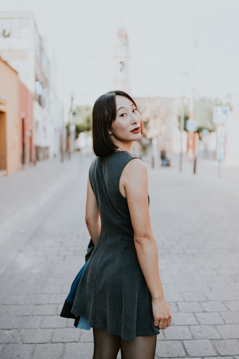 Woman in a gray dress looking back on a sunny, urban street with a classic vibe.