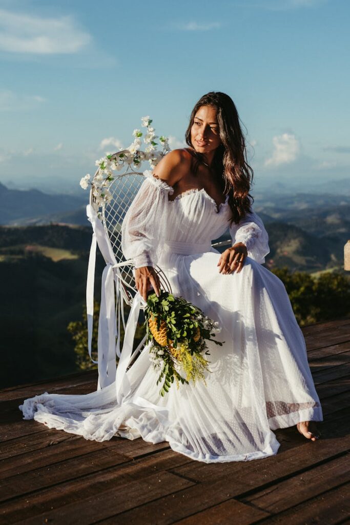 A woman in a flowing white dress sits gracefully on a decorated chair overlooking a breathtaking mountain landscape.