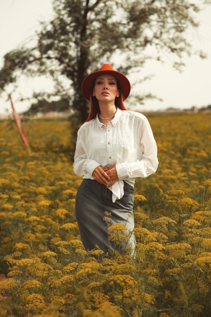 Woman in a field of yellow flowers wearing a stylish hat and white blouse, embodying elegance.