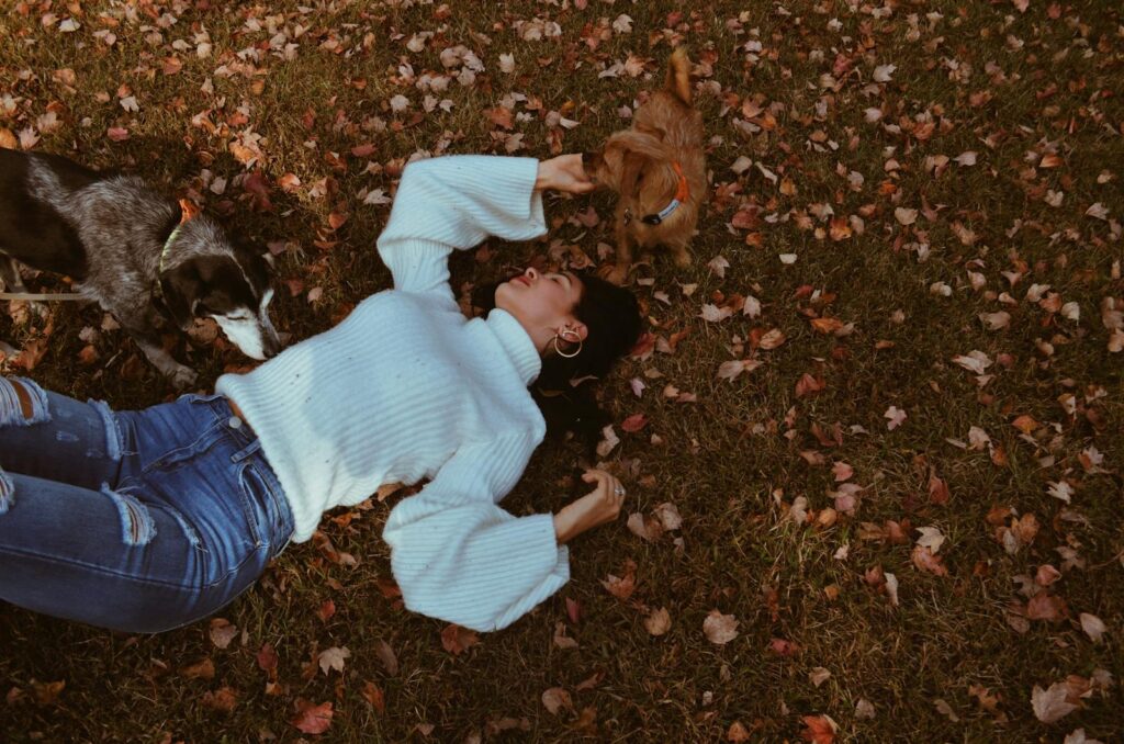 Woman lying on grass with dogs amidst fall leaves, enjoying outdoor autumn day.