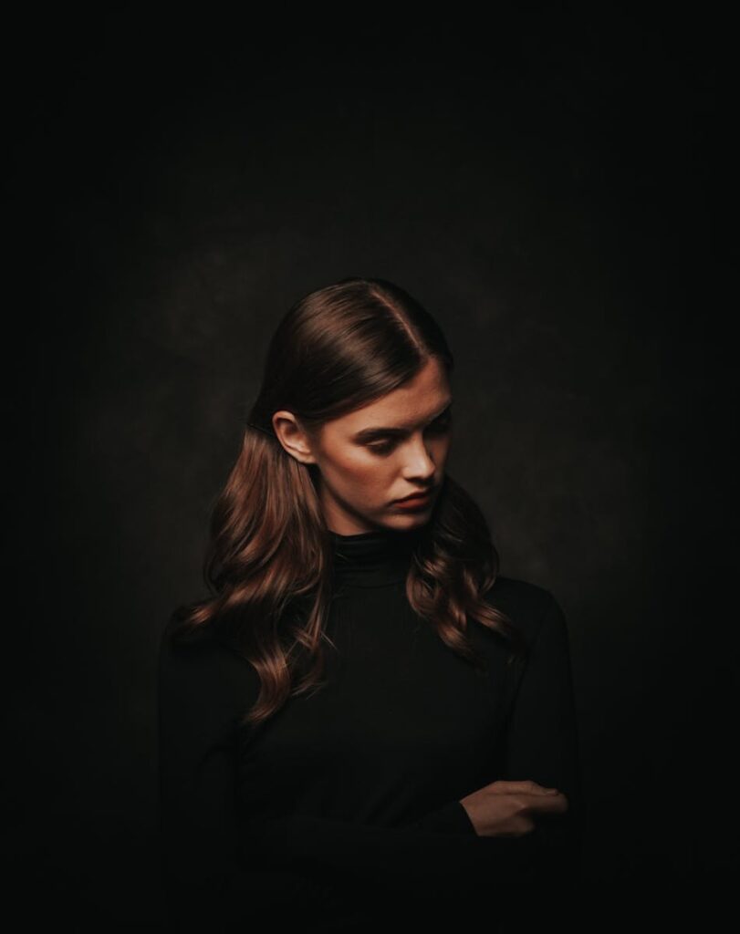 A moody portrait of a woman looking down pensively, set against a dark background.
