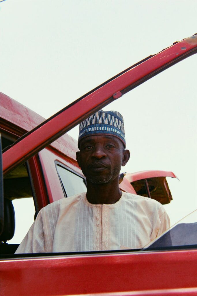 A man wearing traditional attire stands next to a red car, displaying a thoughtful expression.