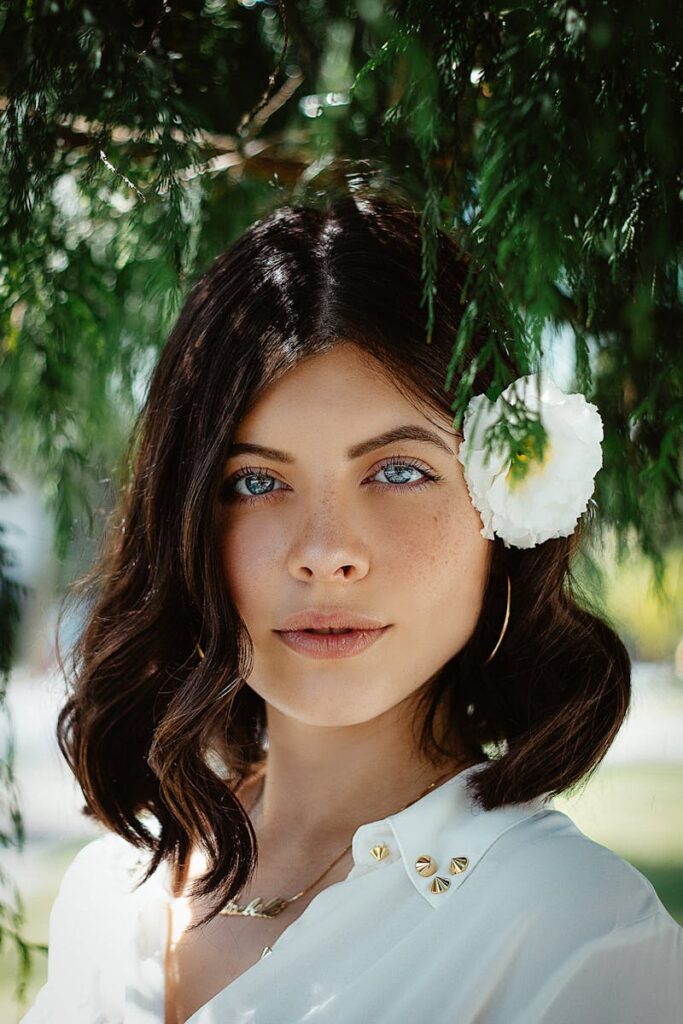 Close-up portrait of a woman with blue eyes and dark hair wearing a white flower outdoors.