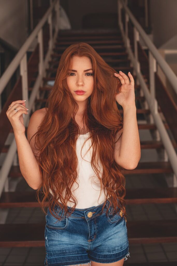 A stylish woman with long red hair poses on stairs in casual attire.
