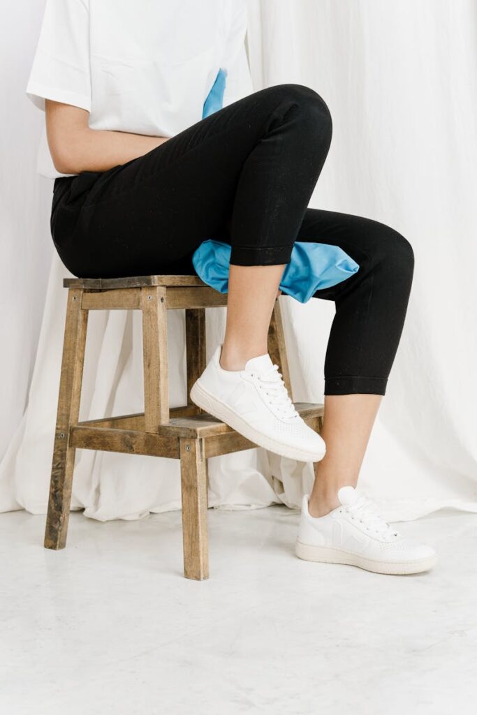 Person sitting on a wooden stool wearing black leggings and white sneakers against a white background.