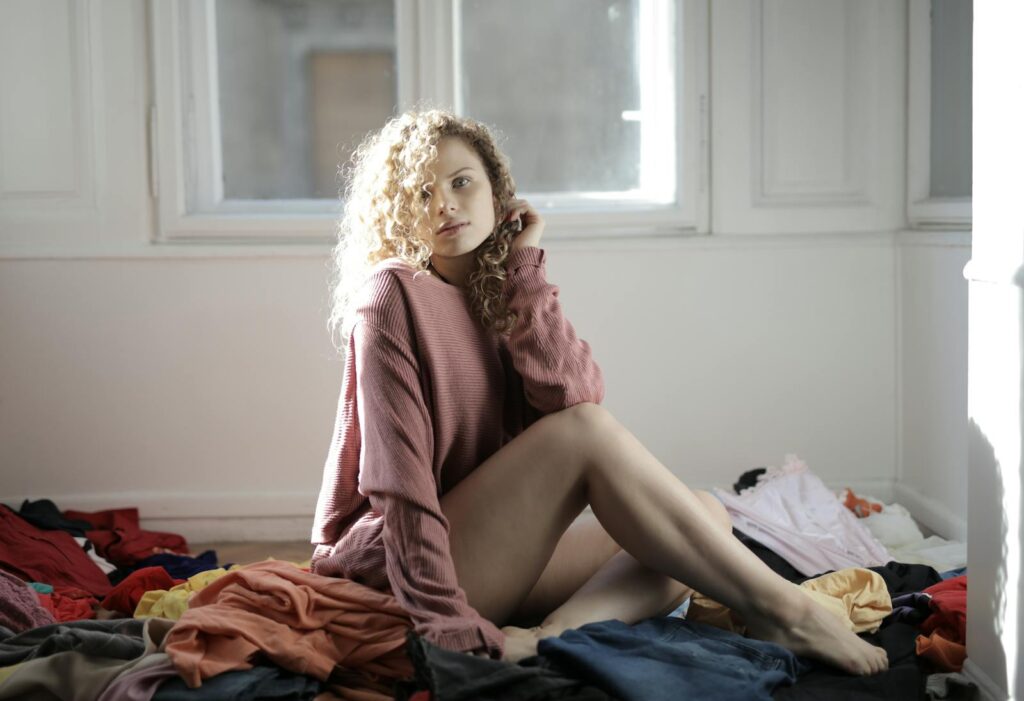 Young woman in a pink sweater sitting amidst a pile of clothes in a bright room.