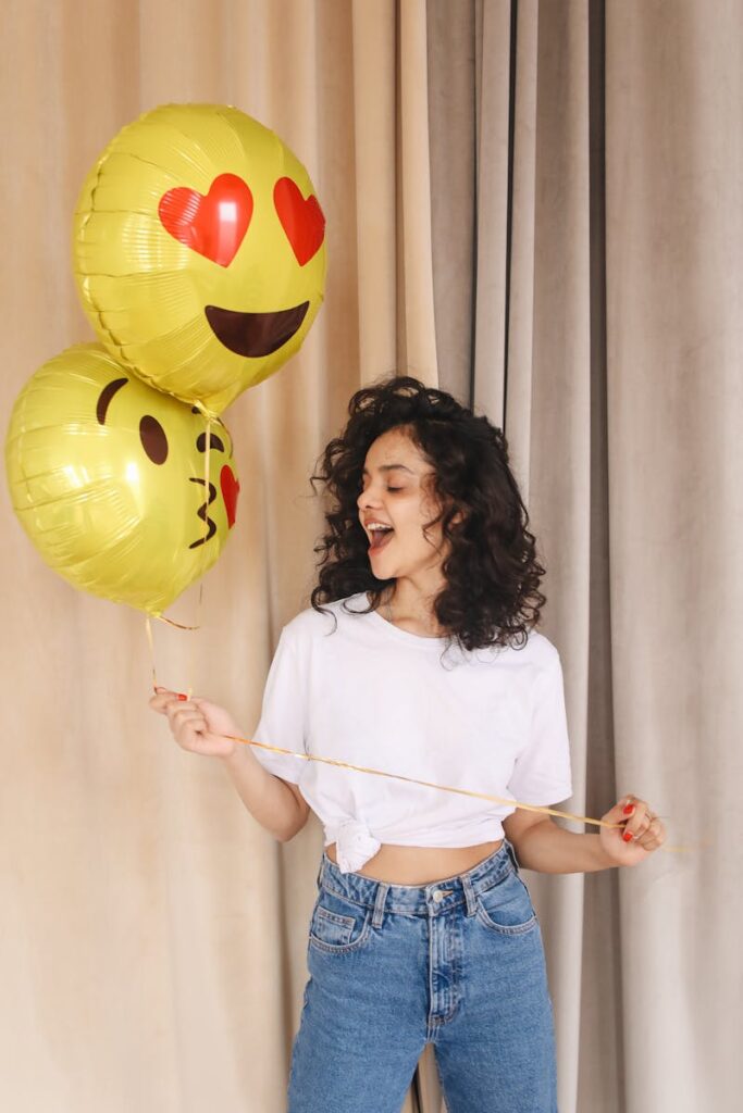 Cheerful woman with curly hair holding emoji balloons indoors, expressing joy and celebration.