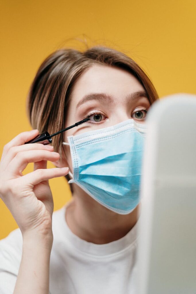 Portrait of a woman applying mascara while wearing a face mask.