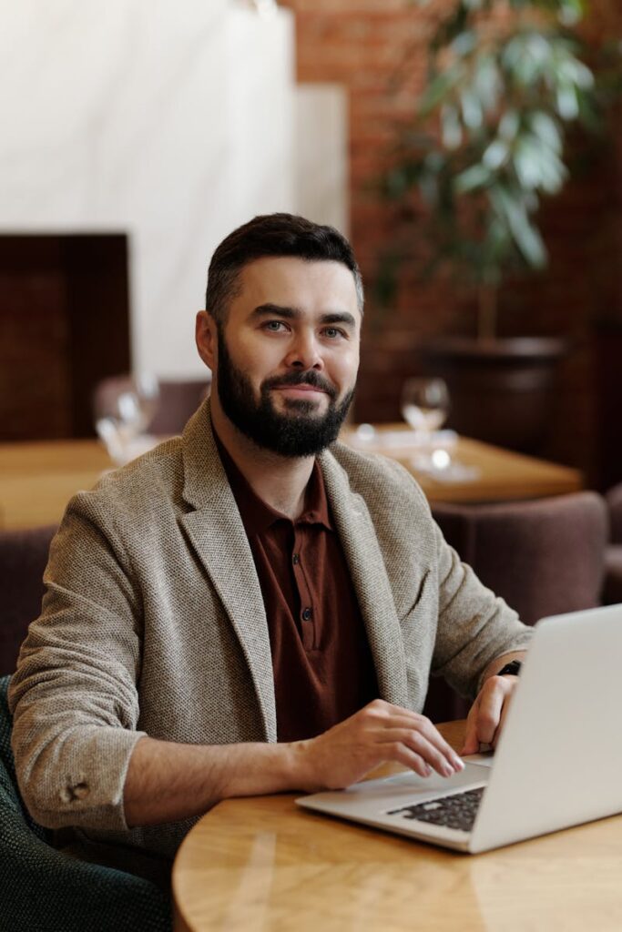 Businessman with a beard and blazer using a laptop in a stylish cafeteria setting.