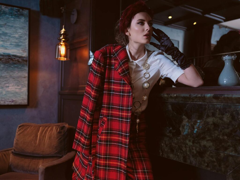 A stylish woman wearing a red tartan coat, leaning gracefully indoors.
