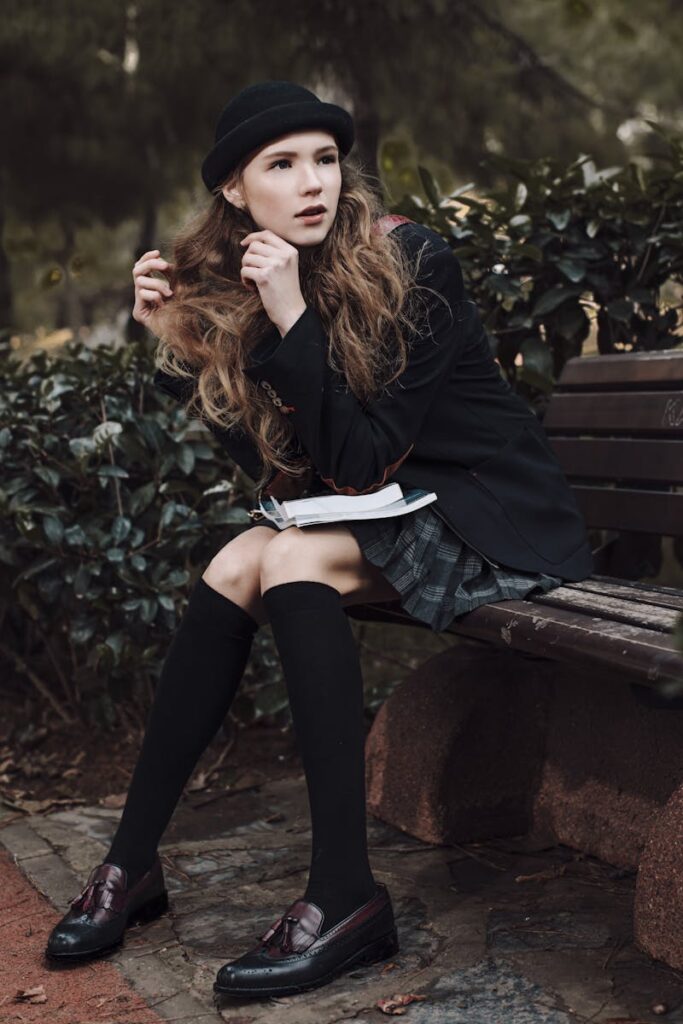 Stylish woman in dark academia attire seated on a park bench with thoughtful look, surrounded by nature.