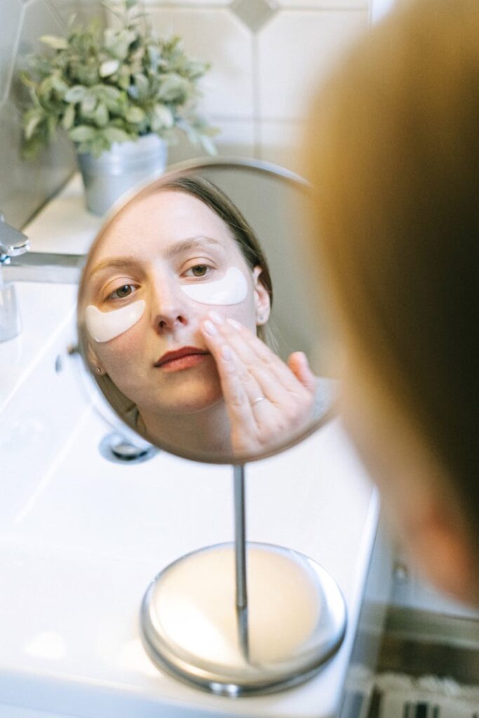 Woman applying eye patches as part of her skincare routine, reflected in bathroom mirror.