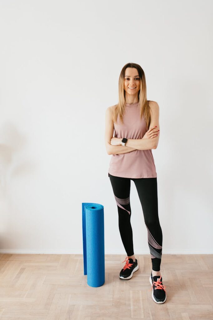 Full body female athlete wearing sportswear and sneakers standing against white wall with crossed arms near blue sport mat before yoga practice and looking at camera with smile