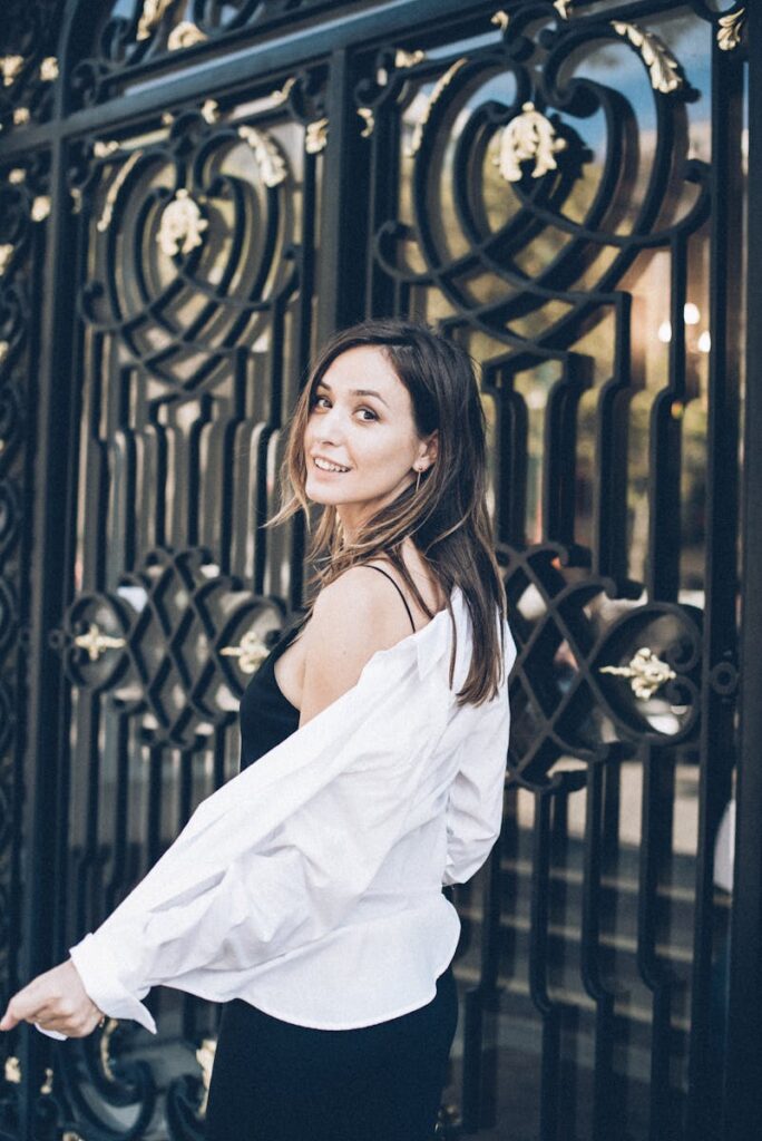 Stylish woman in a black dress poses by an ornate gate, exuding elegance and charm.