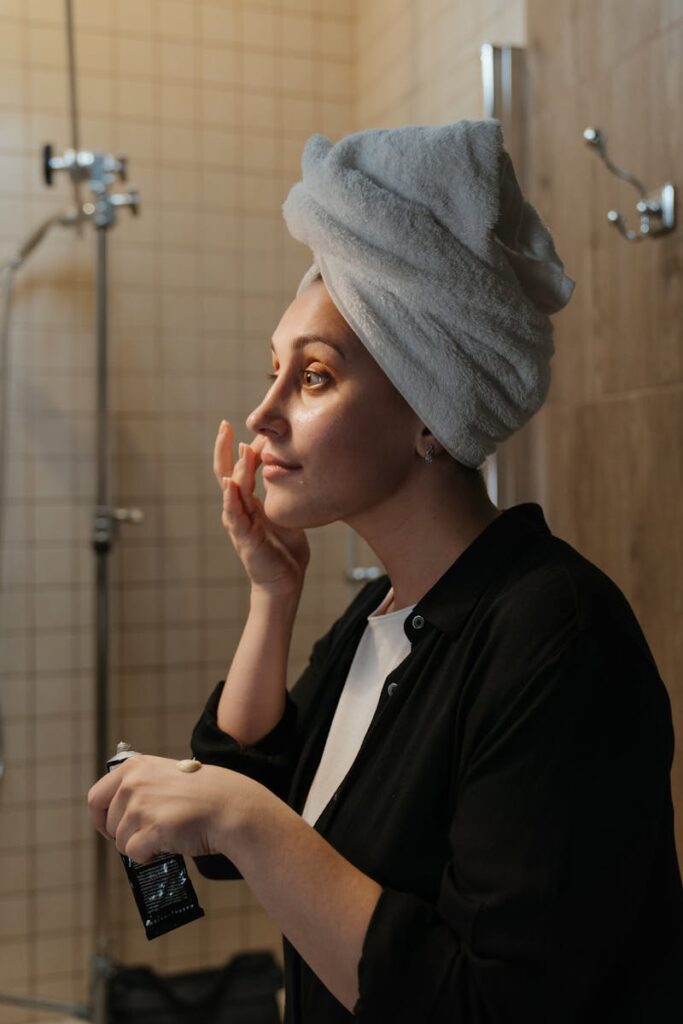 A woman in a towel applies skincare cream in her bathroom during a self-care routine.