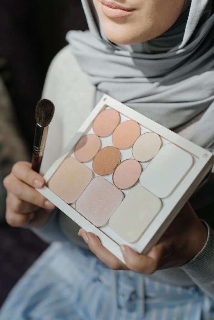 Woman in hijab applying makeup with an eyeshadow palette under natural lighting.