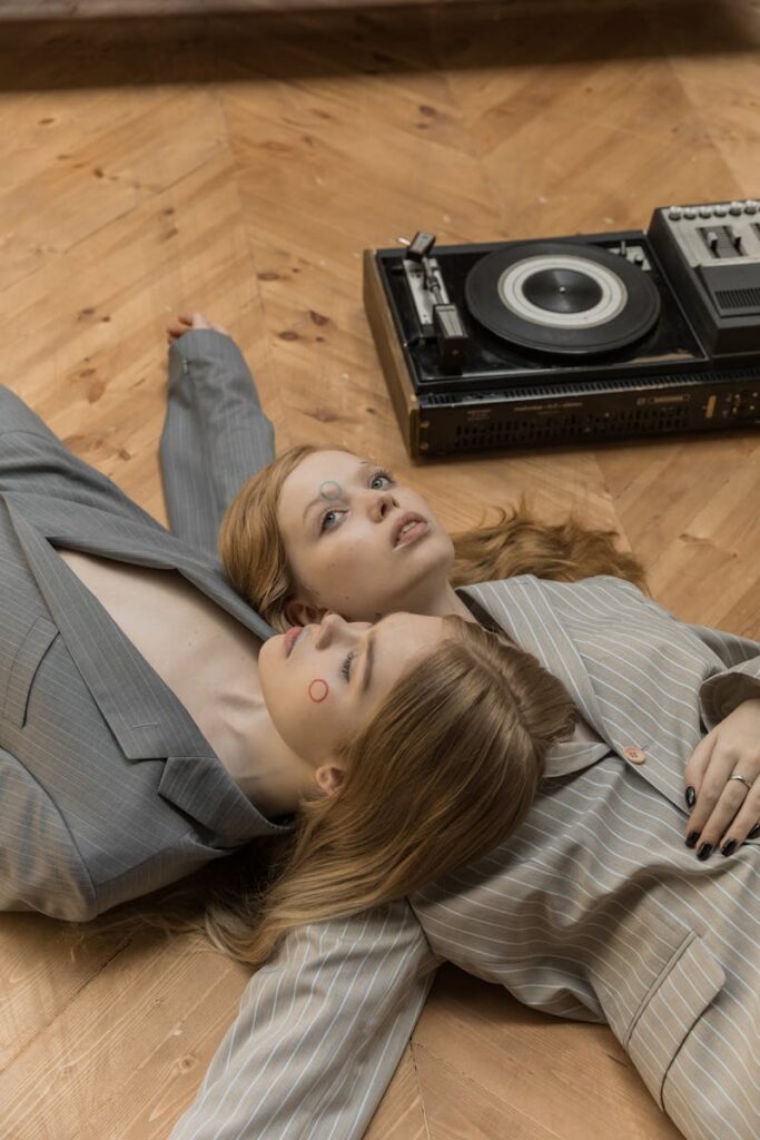 Stylish portrait of two women lying on wooden floor with a vintage record player, showcasing fashion and music themes.