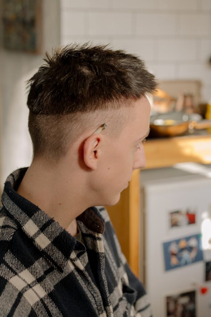 A young man with a distinctive undercut hairstyle sits indoors, wearing a checkered shirt.