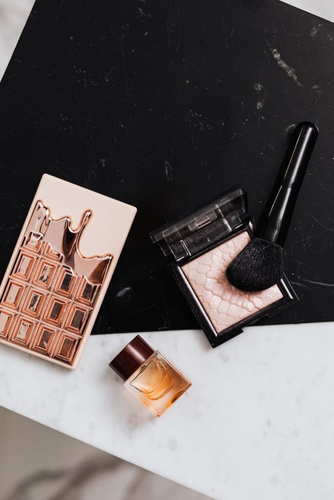 Top view of luxury makeup items including palette, highlighter, and perfume bottle on a marble surface.