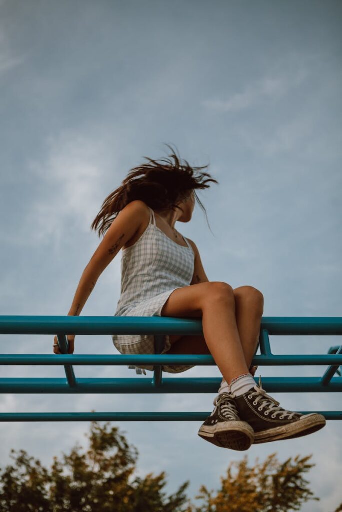A young woman with flowing hair and sneakers sits on blue monkey bars against a summer sky.