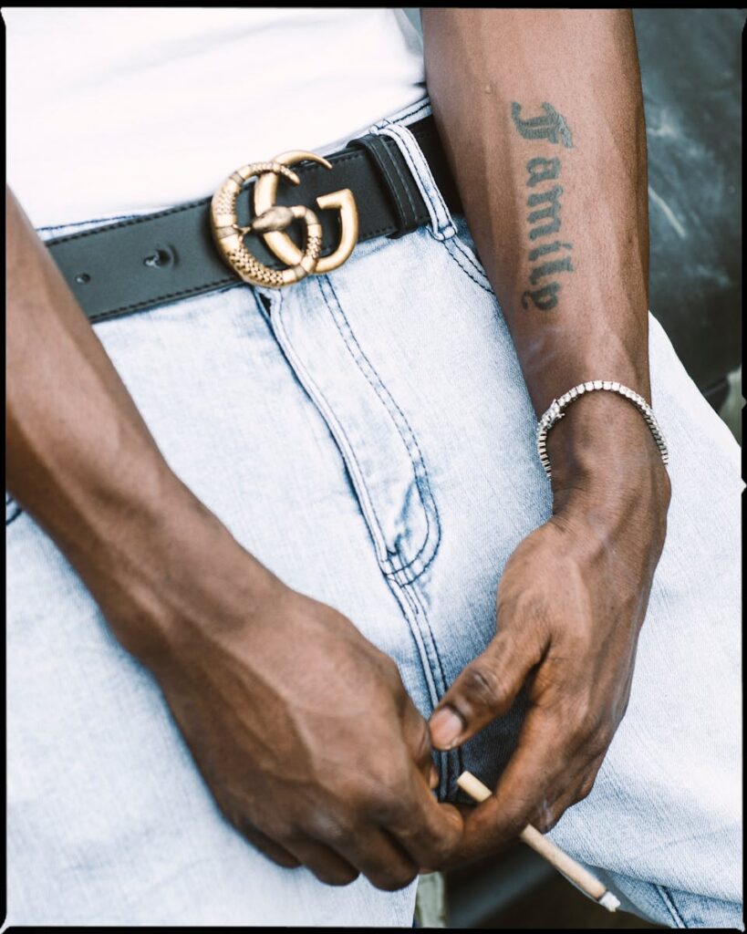 Close-up of a man's tattooed hands with a luxury belt, holding a cigarette.