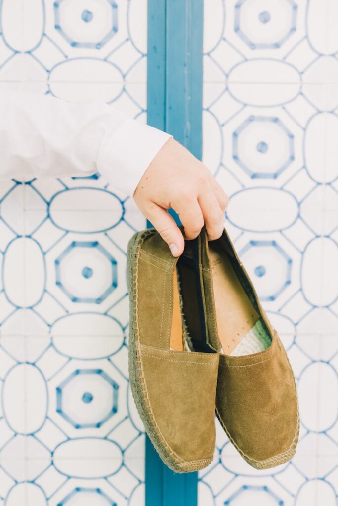A hand holding brown suede espadrilles against a decorative blue and white tile background.