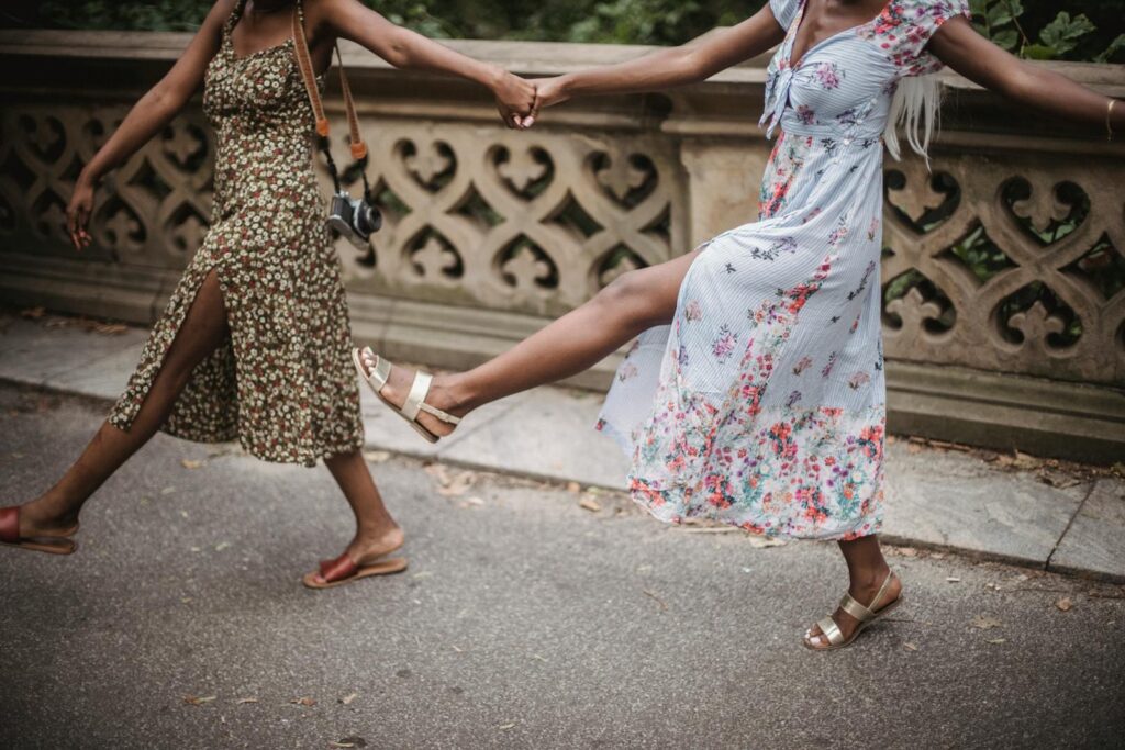 Two women dance in floral dresses while holding hands, capturing a joyful outdoor moment.