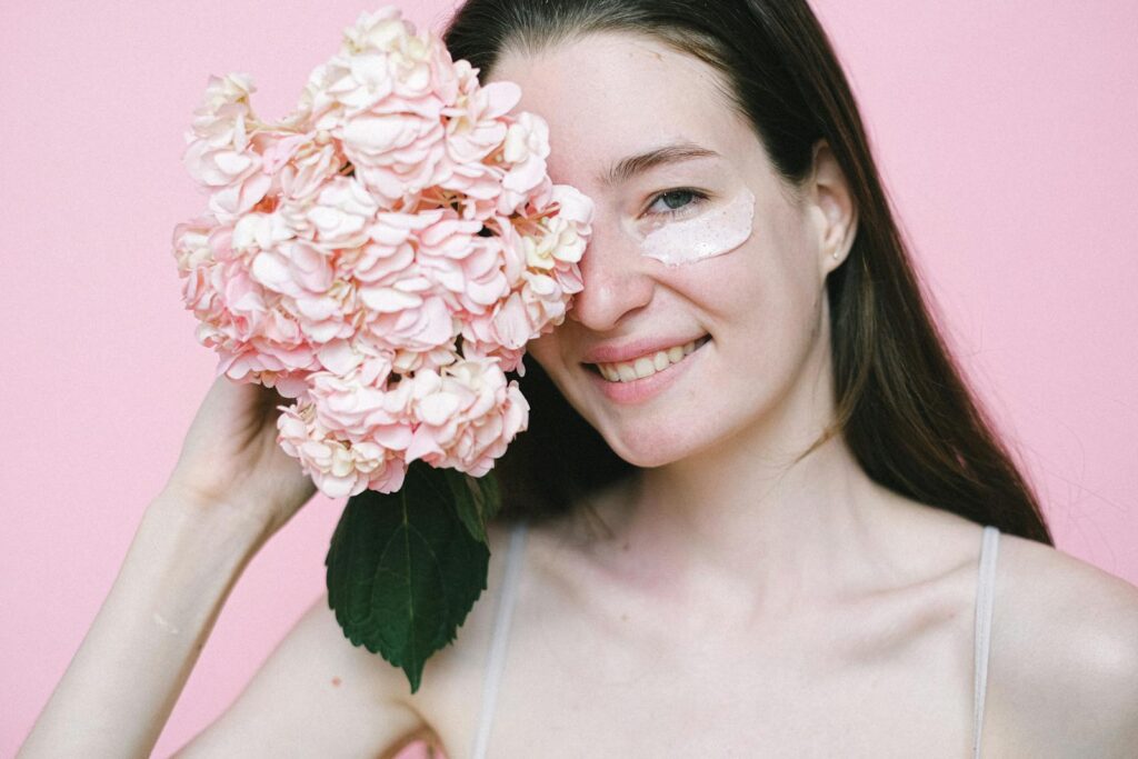 Cheerful woman with skincare cream and hydrangea against pink background, conveying natural beauty.