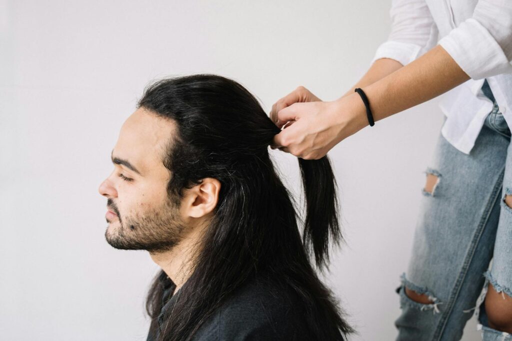 Profile view of a man with long hair being styled by a hairdresser indoors.