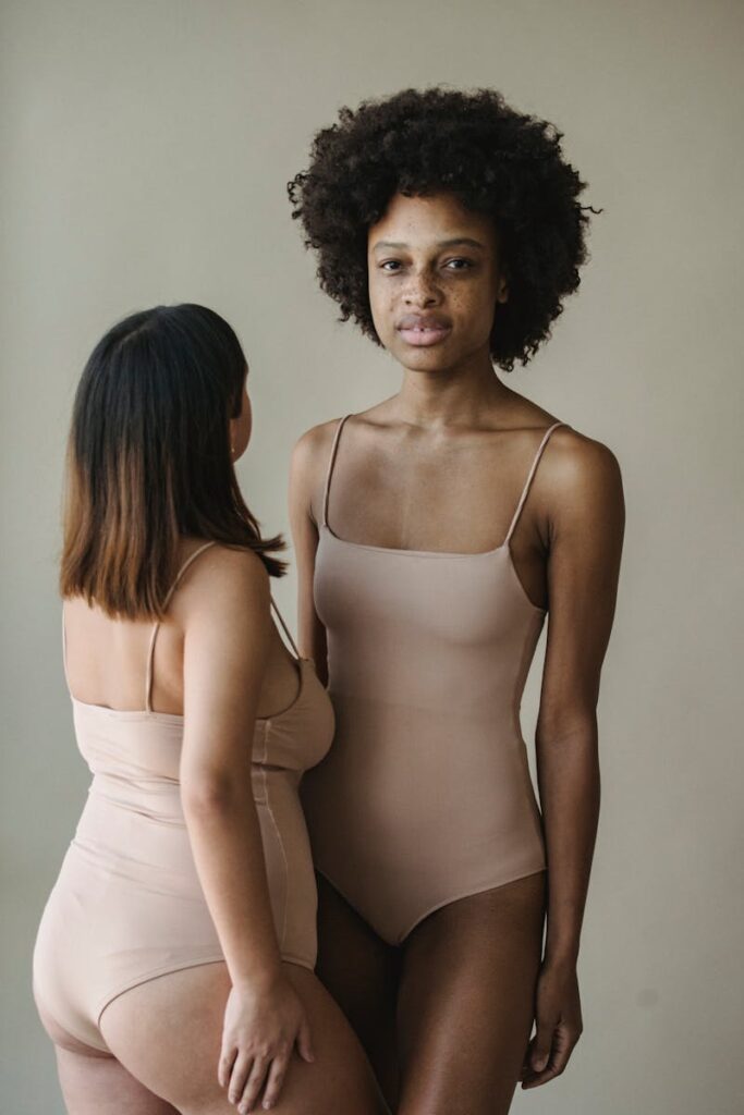 Two women in nude bodysuits standing confidently indoors, showcasing diversity.