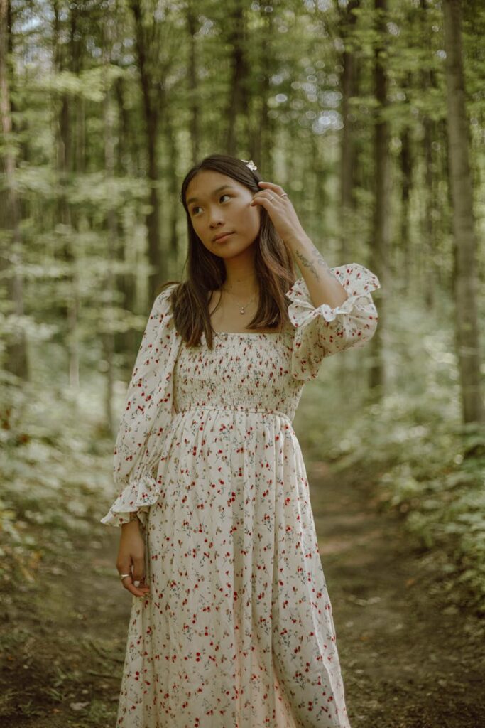 Dreamy young ethnic lady in feminine dress touching long dark hair and looking away while relaxing in green forest on sunny day